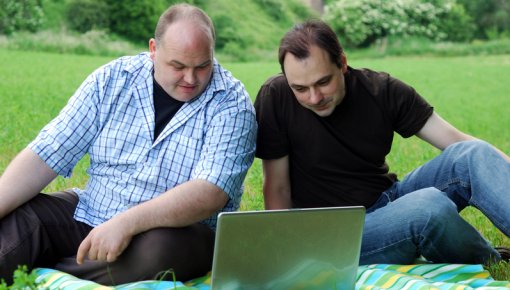Two men sitting outside in a park in front of a laptop 